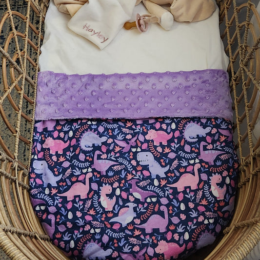 A purple baby blanket with a dinosaur pattern, minky backing, displayed on a wicker surface. handmade by hayley james